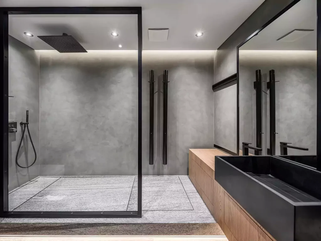 Bathroom in a modern style with gray tiled walls. There is a shower with a glass partition, wooden stand with a black sink and a faucet, large mirror, luminous lamps. Horizontal.